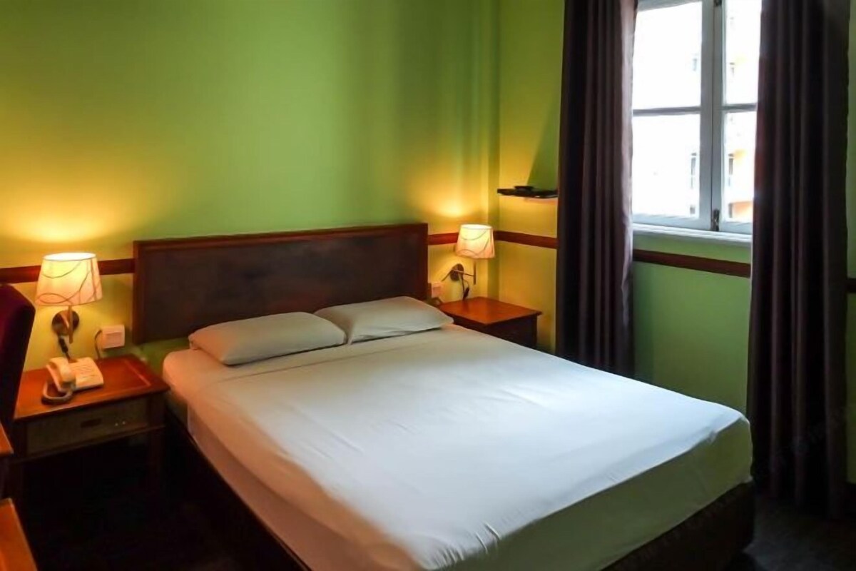 Habitació d'hotel amb un llit individual, dues taules de nit, una tauleta, una làmpada i una finestra amb cortines.