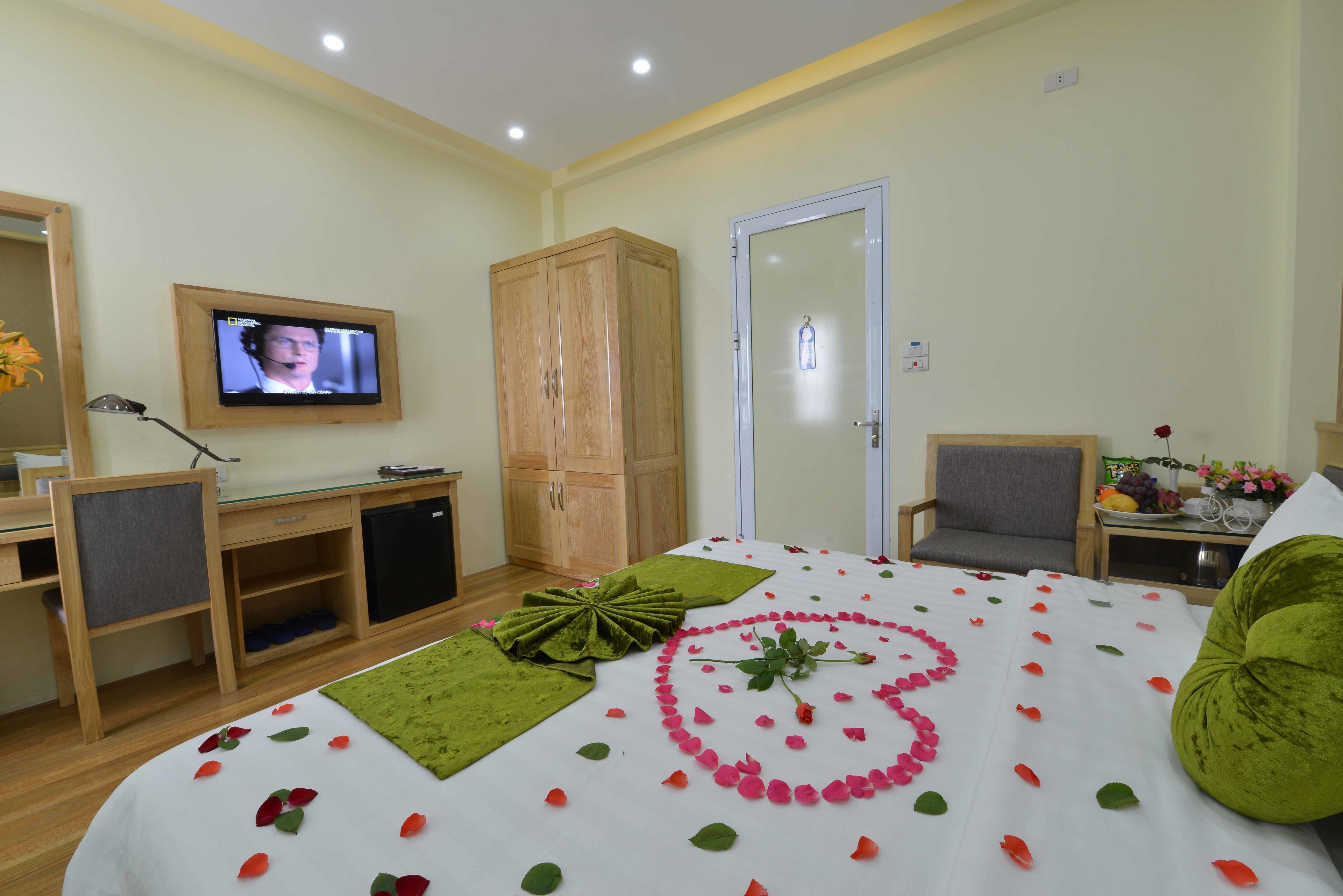 A hotel room with a bed adorned with rose petals and a heart-shaped arrangement, a television mounted on the wall, a desk with a computer, and a wardrobe.