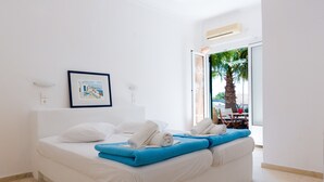 A modern hotel room with a large bed, white bedding, a framed picture on the wall, and a view of palm trees outside. - Memento Perissa (Santorini)