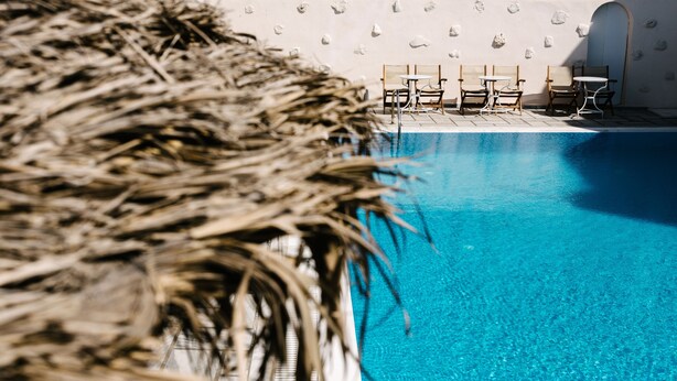 A clear blue pool with lounge chairs and a thatched roof structure. - Memento Perissa (Santorini)