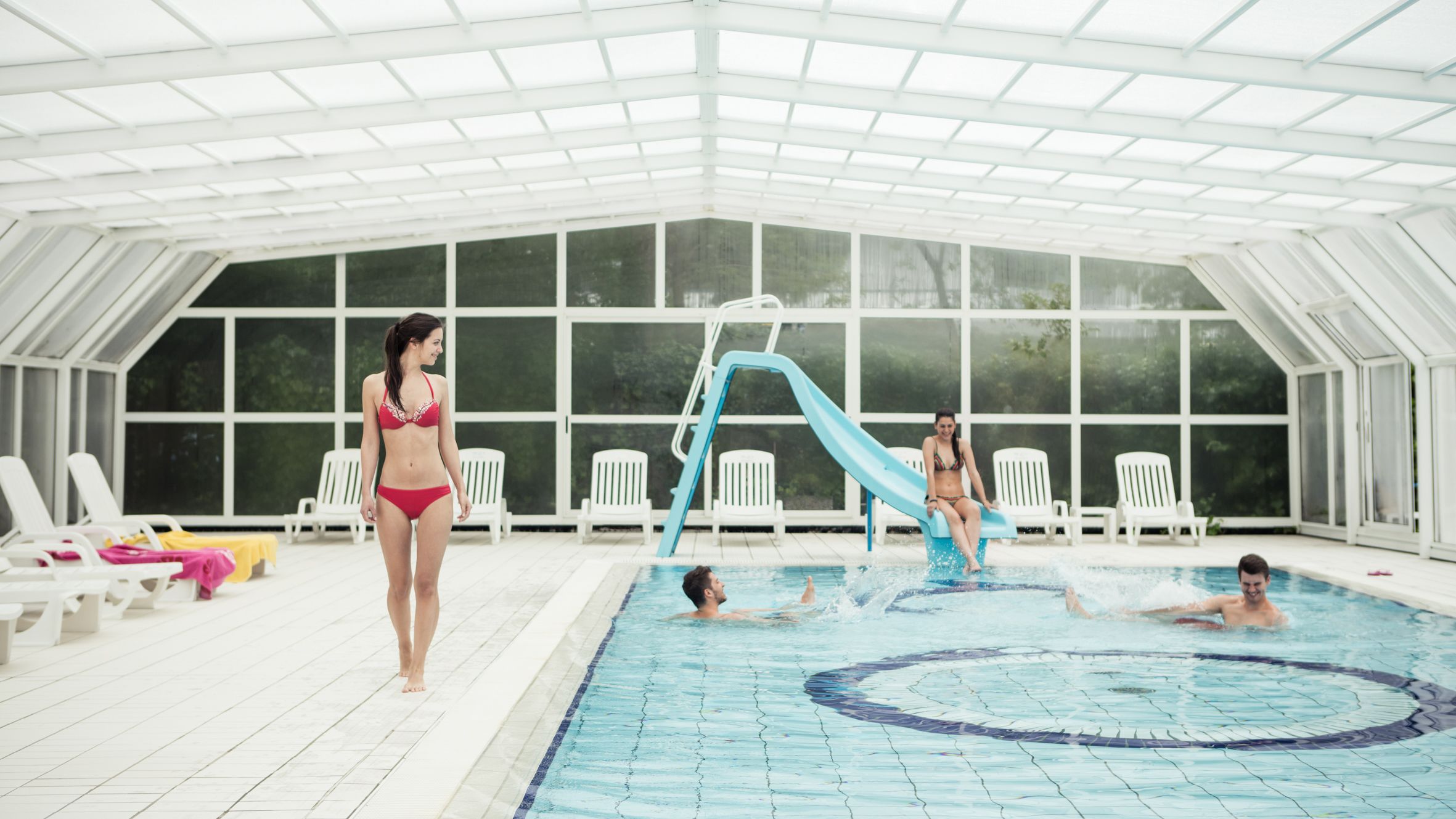En indendørs swimmingpool med vandrutsjebane, liggestole og en stor væg med vinduer.