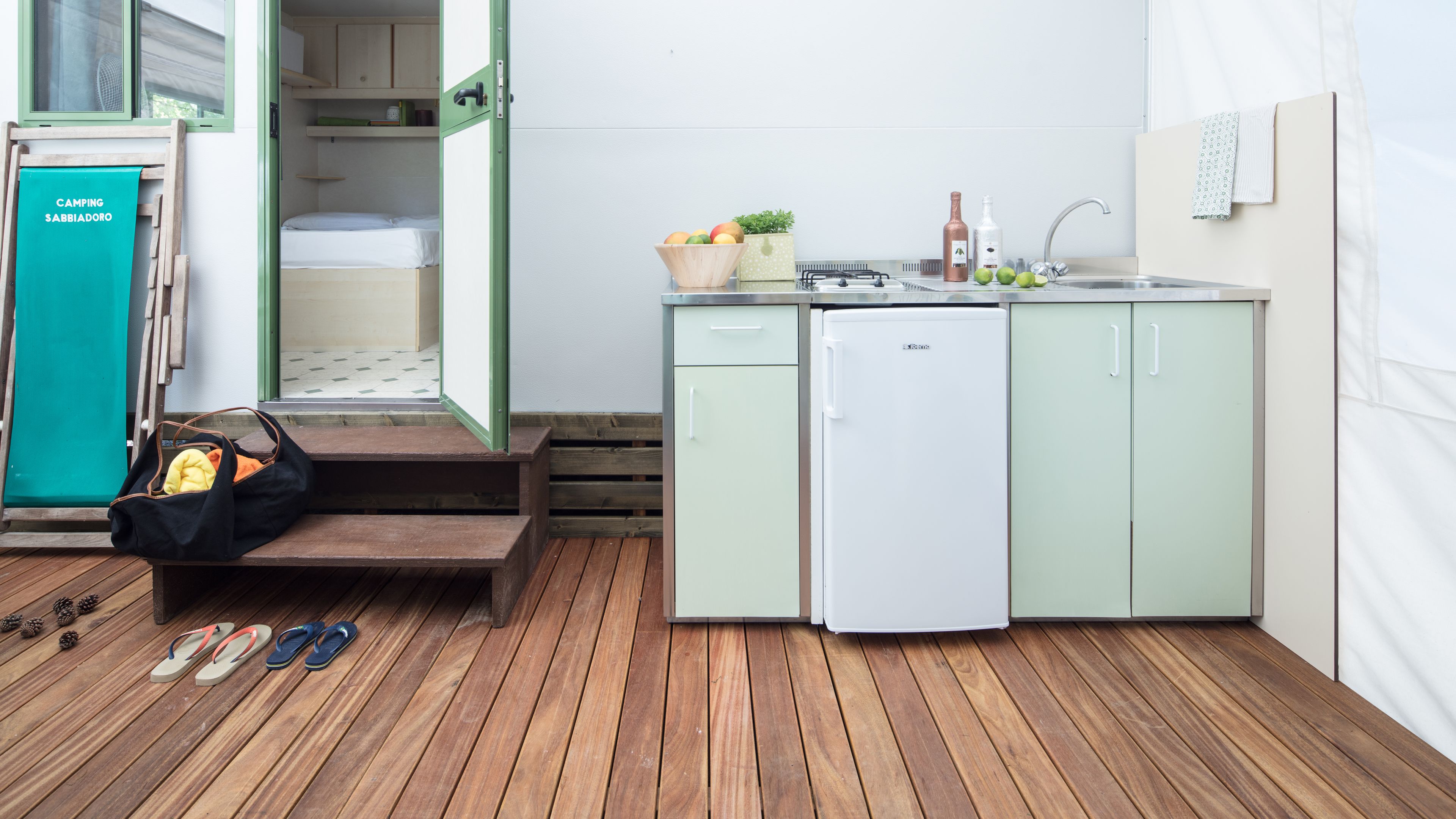 A compact kitchen with a sink, a small refrigerator, and a wooden deck with a bench and sandals.