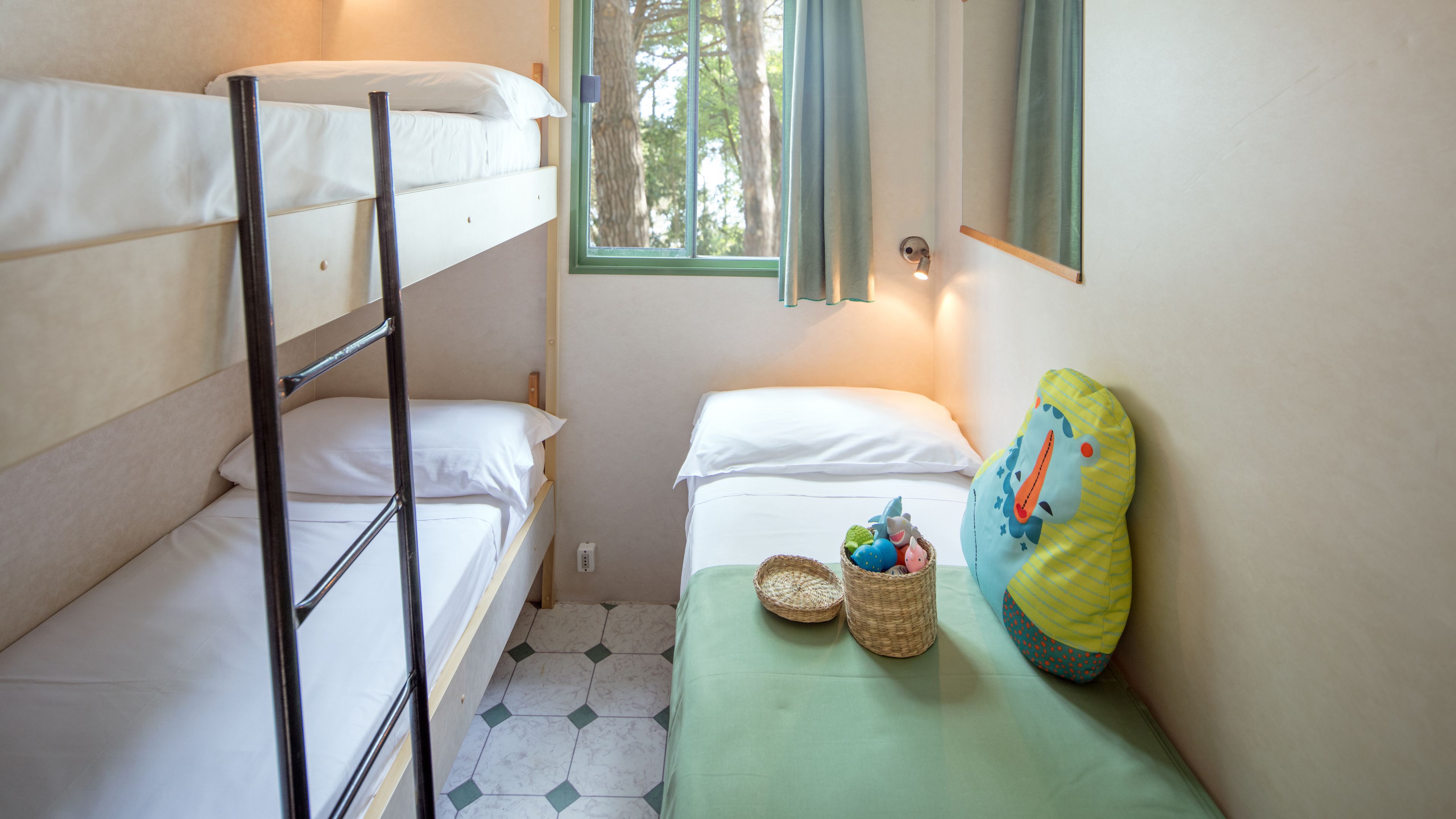Een kleine, compacte kamer met stapelbedden, een enkel bed met een groene sprei en een raam met uitzicht op bomen.