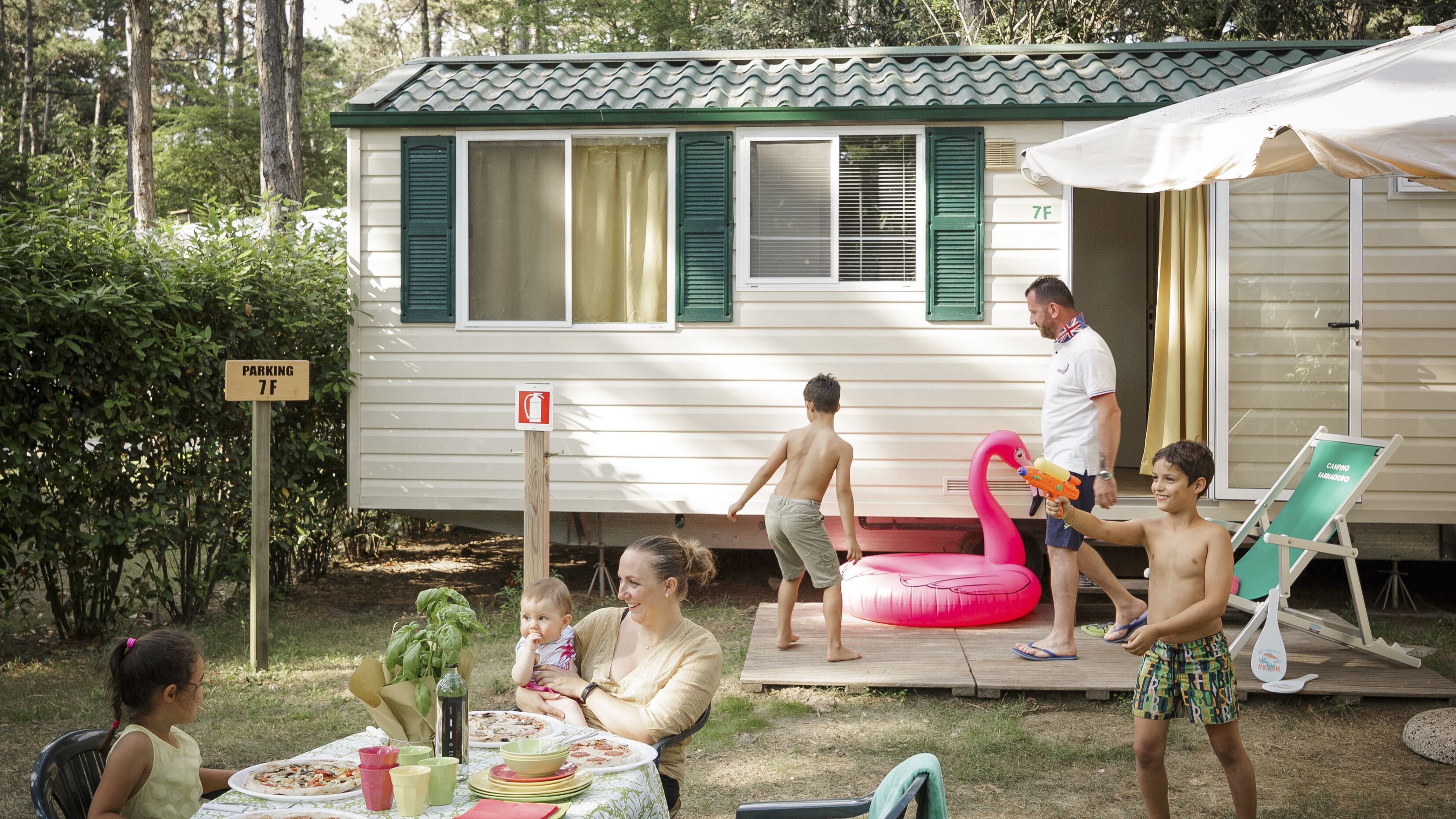 Una casa móvil con un área de patio, una mesa de comedor con comida y un niño jugando con un flotador de flamenco.