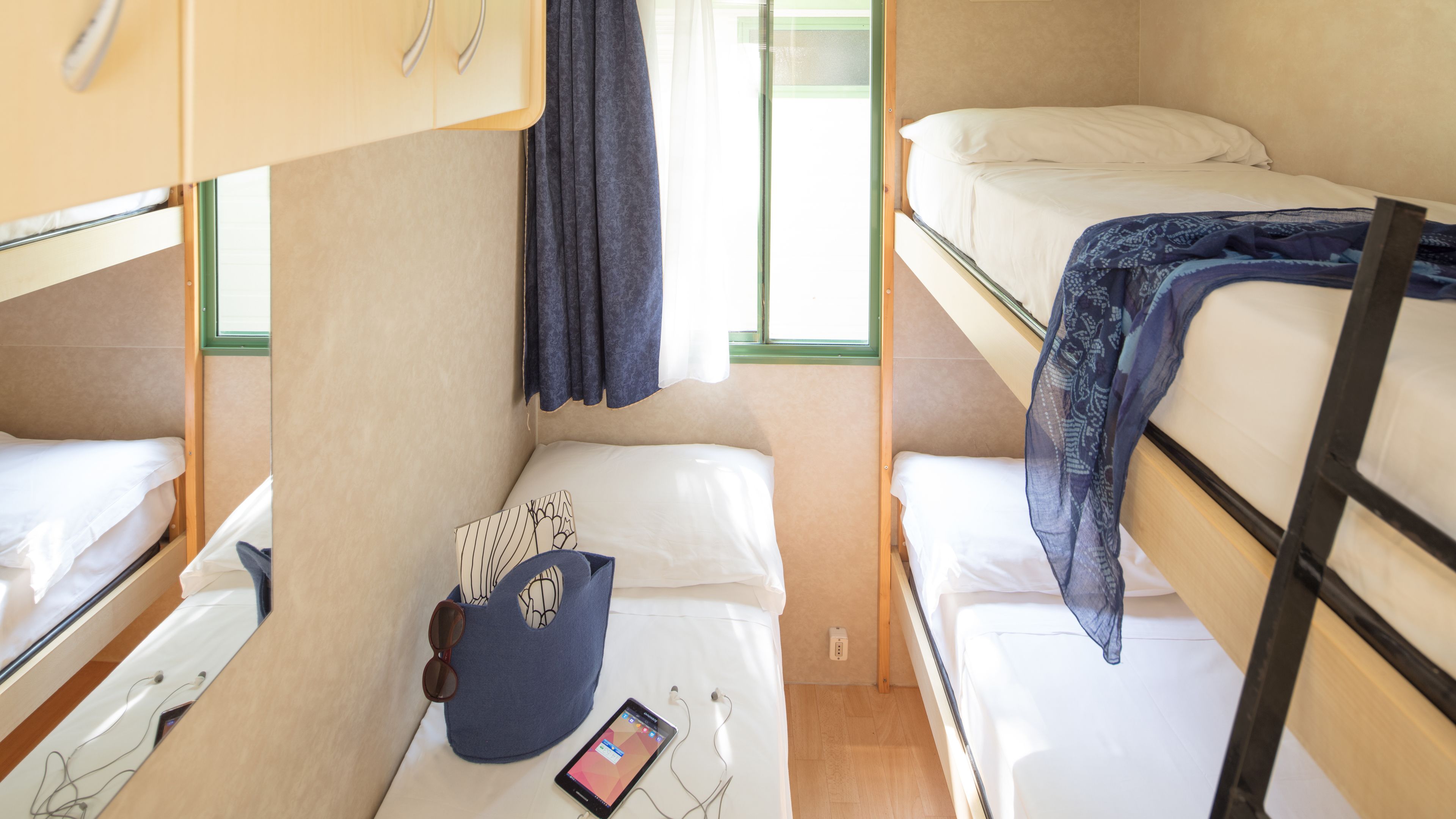 Habitació petita amb literes, una taula on hi ha un telèfon mòbil i una finestra amb cortines.