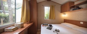 A bedroom with a wooden headboard, a bedside table with books and a camera, a window with curtains, and a shelf with books and a bag. - Camping Sabbiadoro (Lignano Sabbiadoro)