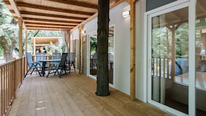 Una terraza de madera con una mesa y sillas, un gran tronco de árbol y una puerta corrediza de vidrio que da a un espacio cubierto.