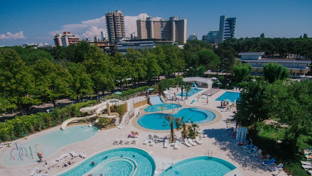 Un extenso complejo de piscinas al aire libre con varias piscinas y un tobogán acuático, rodeado de árboles y sillas de descanso.