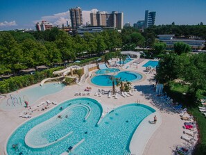 A large outdoor swimming pool complex with multiple pools and a water slide, surrounded by trees and lounge chairs. - Camping Sabbiadoro (Lignano Sabbiadoro)