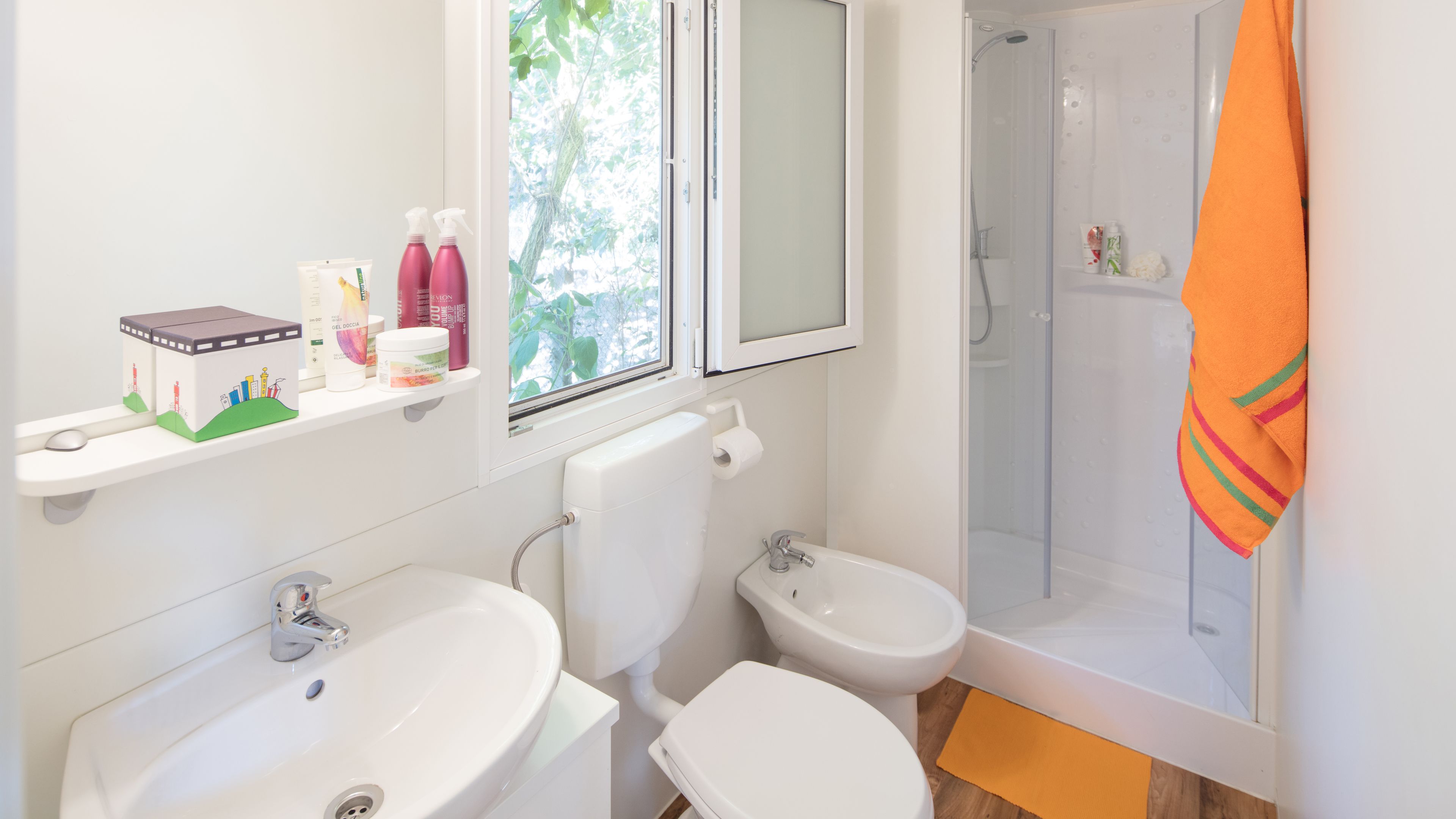 A modern bathroom with two sinks, a toilet, a shower, and a window with a view of greenery.