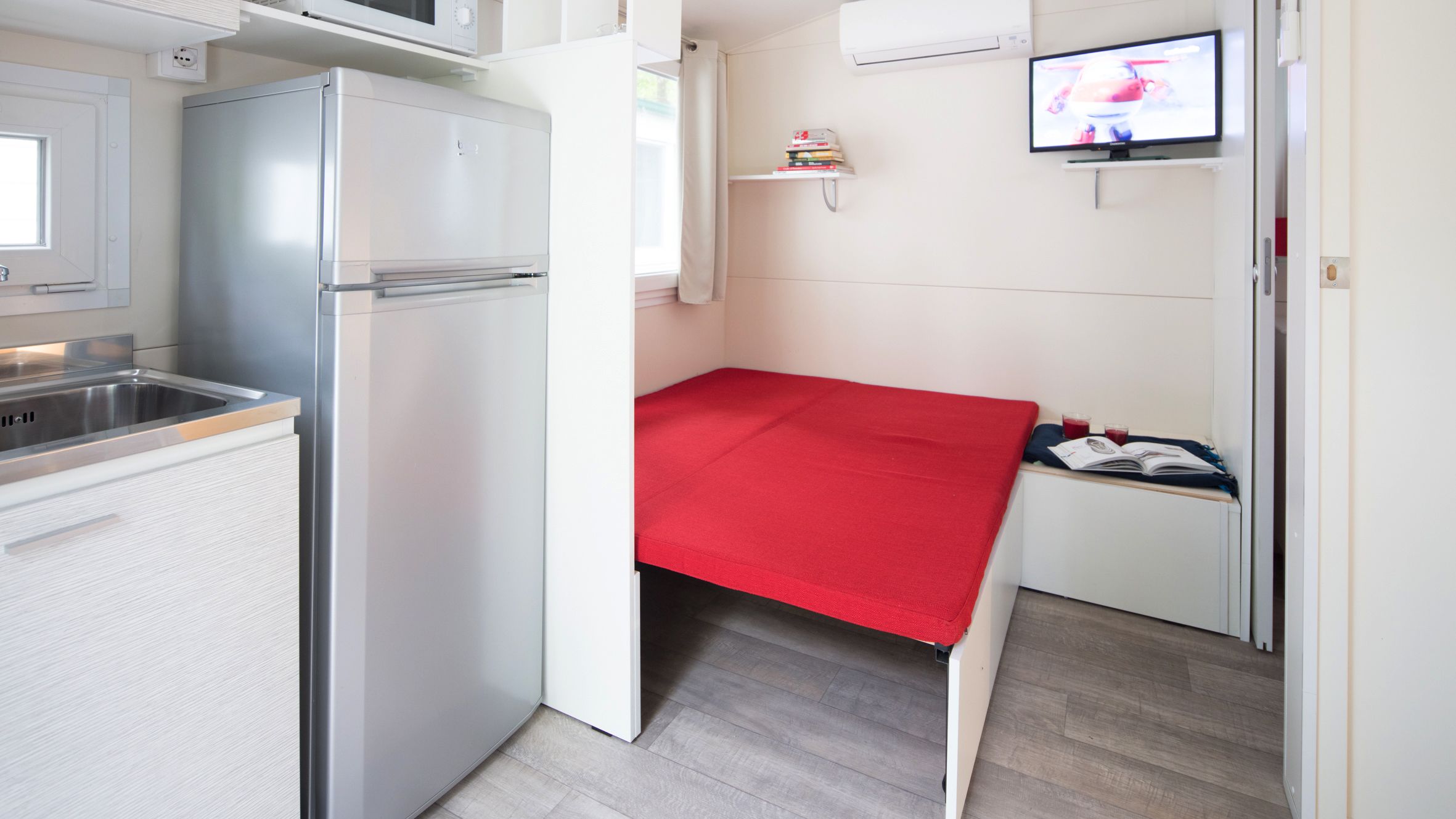 A compact kitchenette with a refrigerator, microwave, and sink. A small TV is mounted on the wall, and a red bedspread is visible.