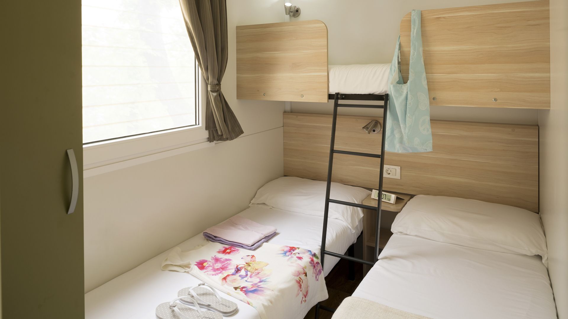 Una habitación pequeña con una cama individual y una litera encima, una ventana con cortinas, una mesita de noche con lámpara y una colcha estampada con motivos florales.