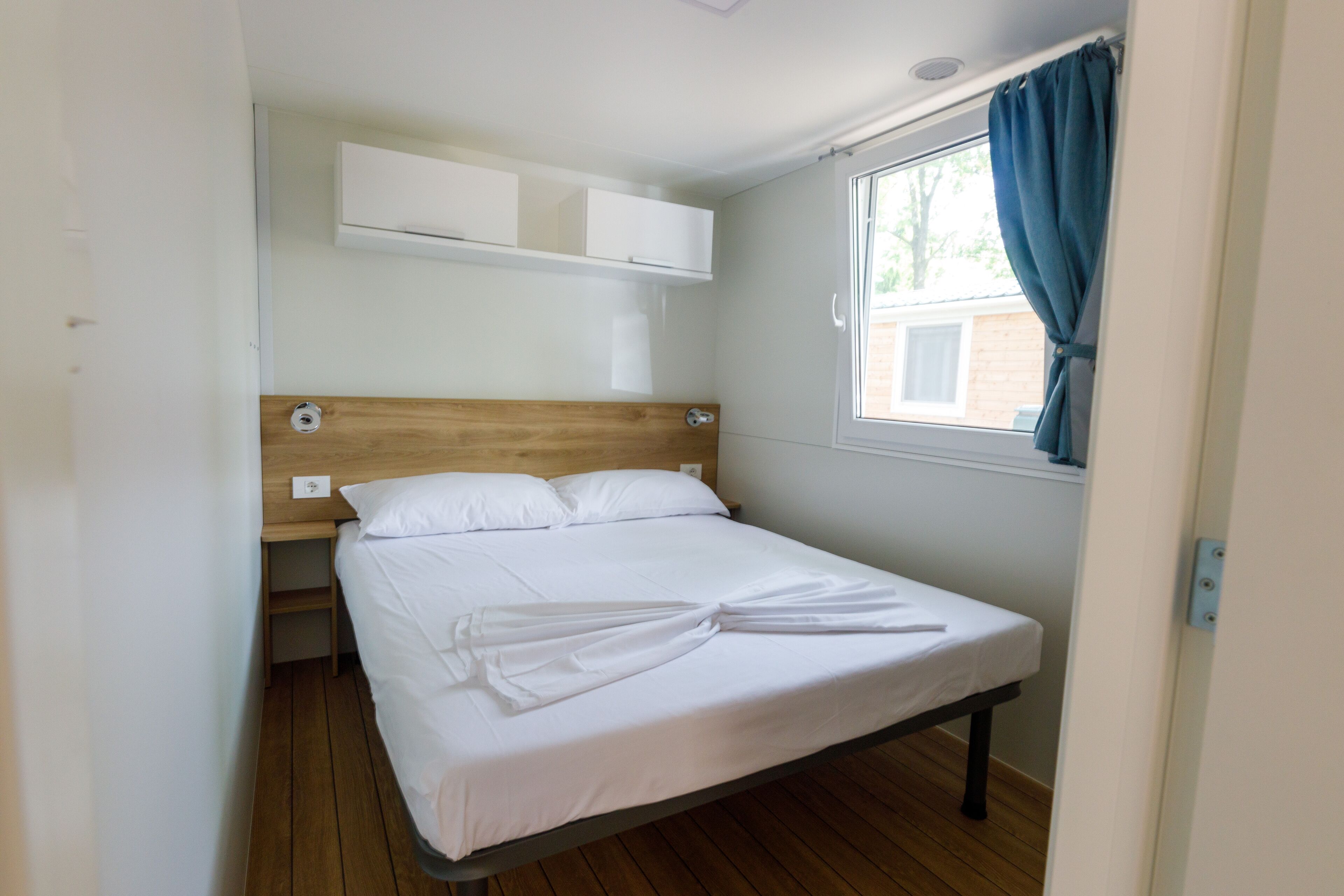 A single bed with a wooden headboard, white bedding, and a window with blue curtains.