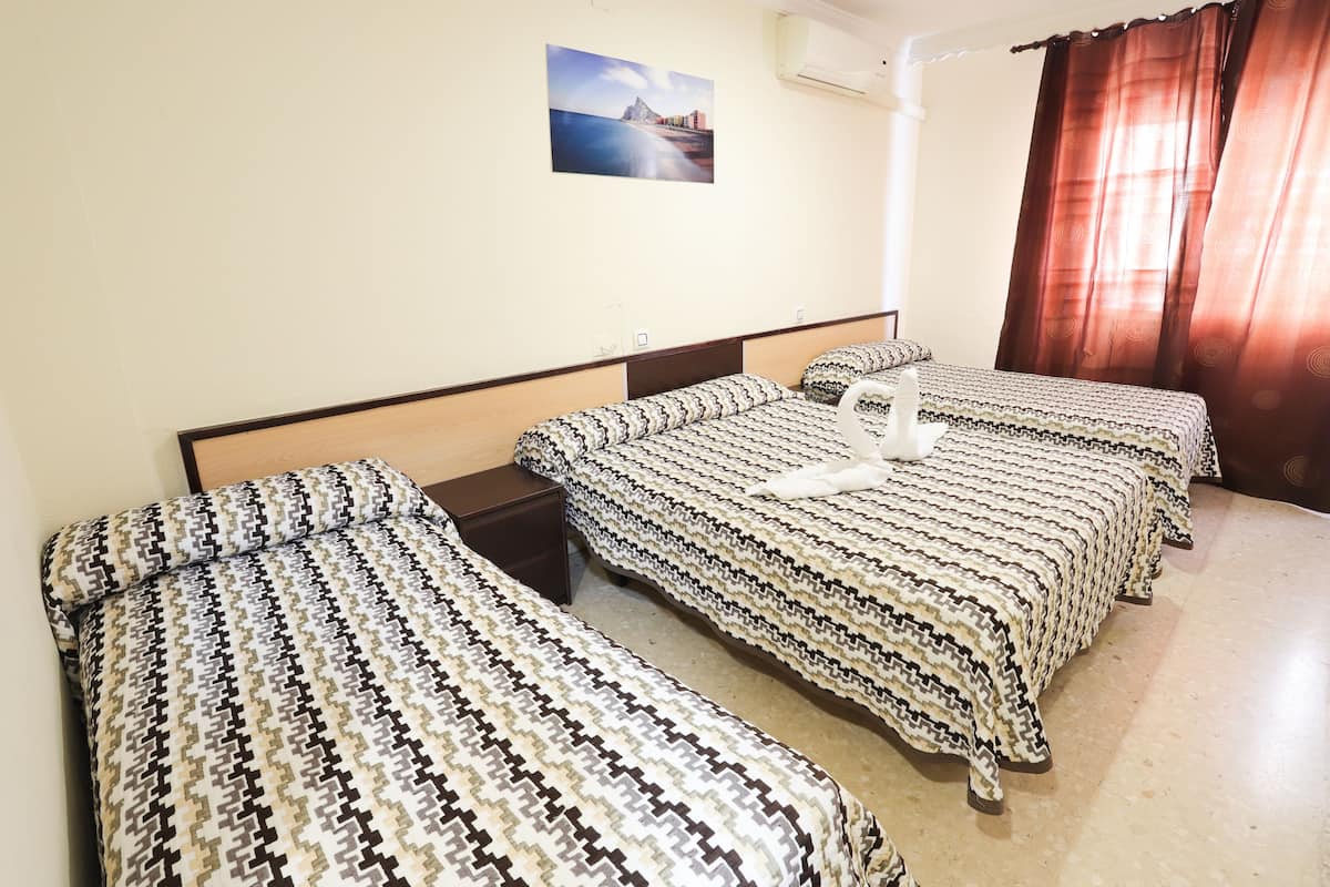 Habitación de hotel con tres camas, cada una con ropa de cama estampada, una mesita de noche, aire acondicionado de pared y un cuadro enmarcado sobre las camas.