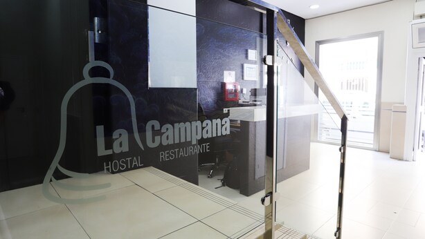 Un hall d’hôtel moderne avec un mur de verre sur lequel est inscrit le nom « La Campana HOSTAL RESTAURANTE ».