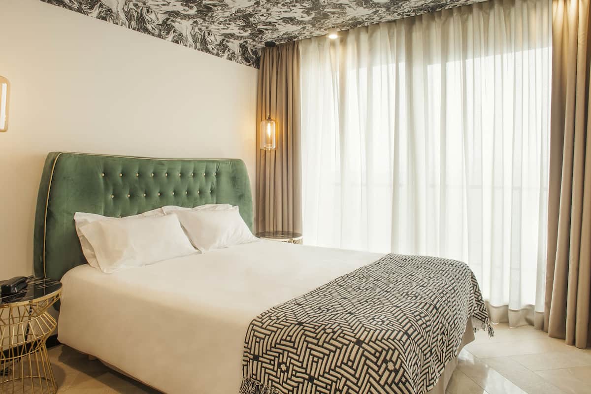 Une chambre d’hôtel avec une tête de lit recouverte d’un tissu vert, un grand lit avec du linge de lit blanc, un plaid à motifs, une table ronde avec un plateau noir et un cadre doré, et un luminaire suspendu.