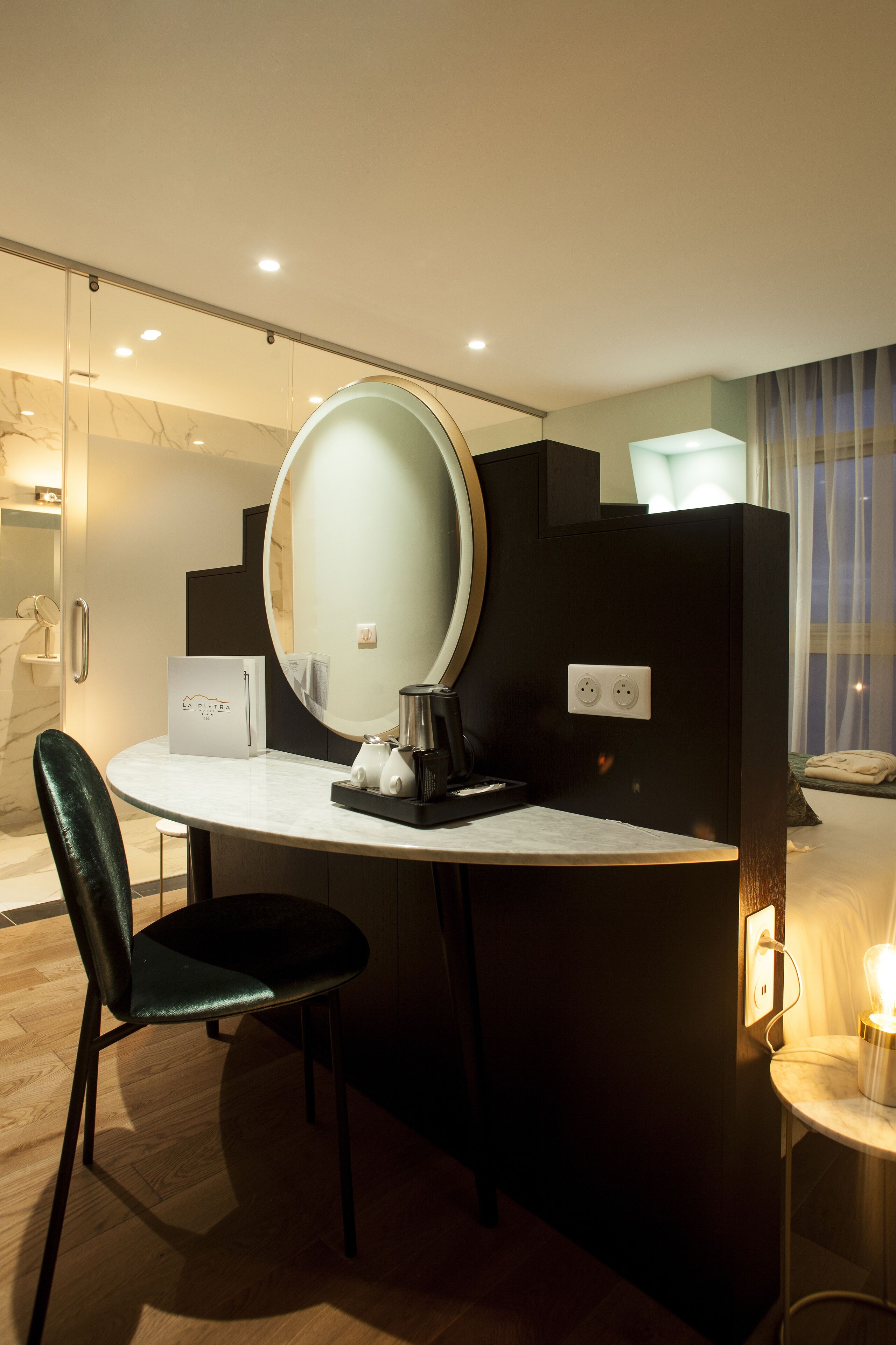 A modern hotel room with a round mirror, a small table with a lamp, a chair, and a minibar.