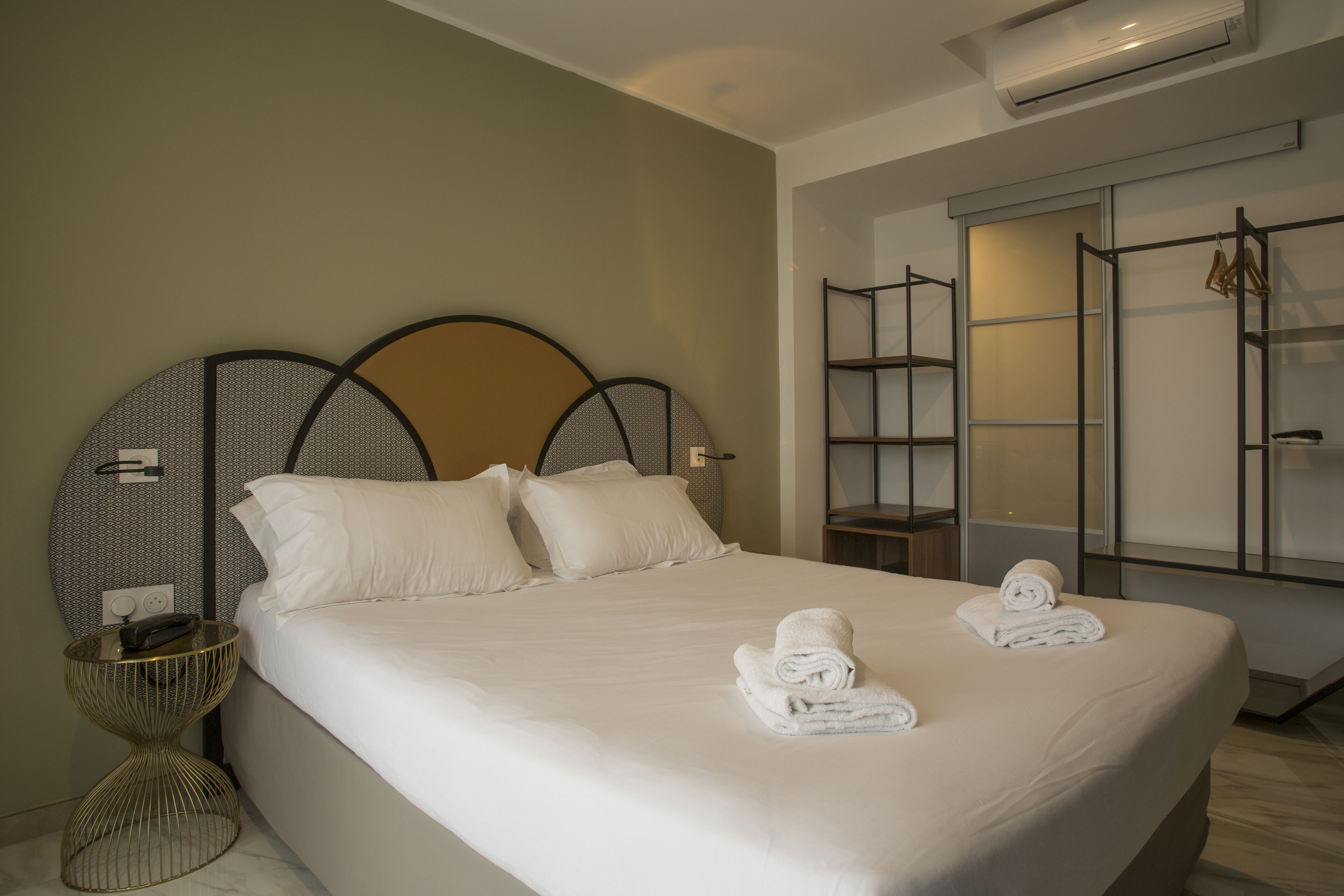 A neatly made bed with white linens and two rolled towels, a bedside table with a lamp, and a wall-mounted shelf with a few items.