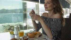 Una persona sentada a una mesa con alimentos para el desayuno, una ventana con vista al mar y un paisaje urbano al fondo.