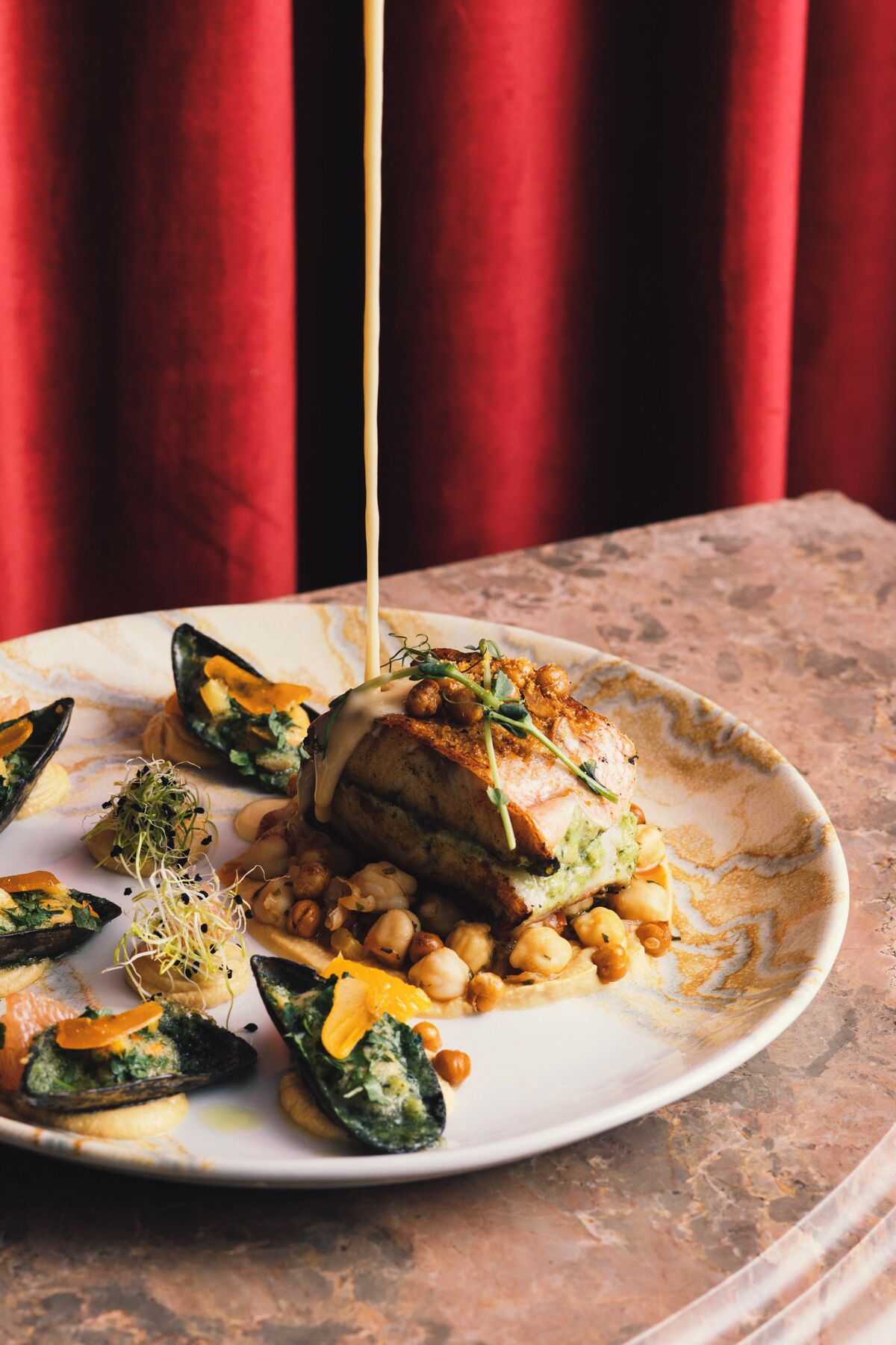 A plate of food with mussels, chickpeas, and a piece of meat, possibly chicken, on a marble table with a red curtain in the background.