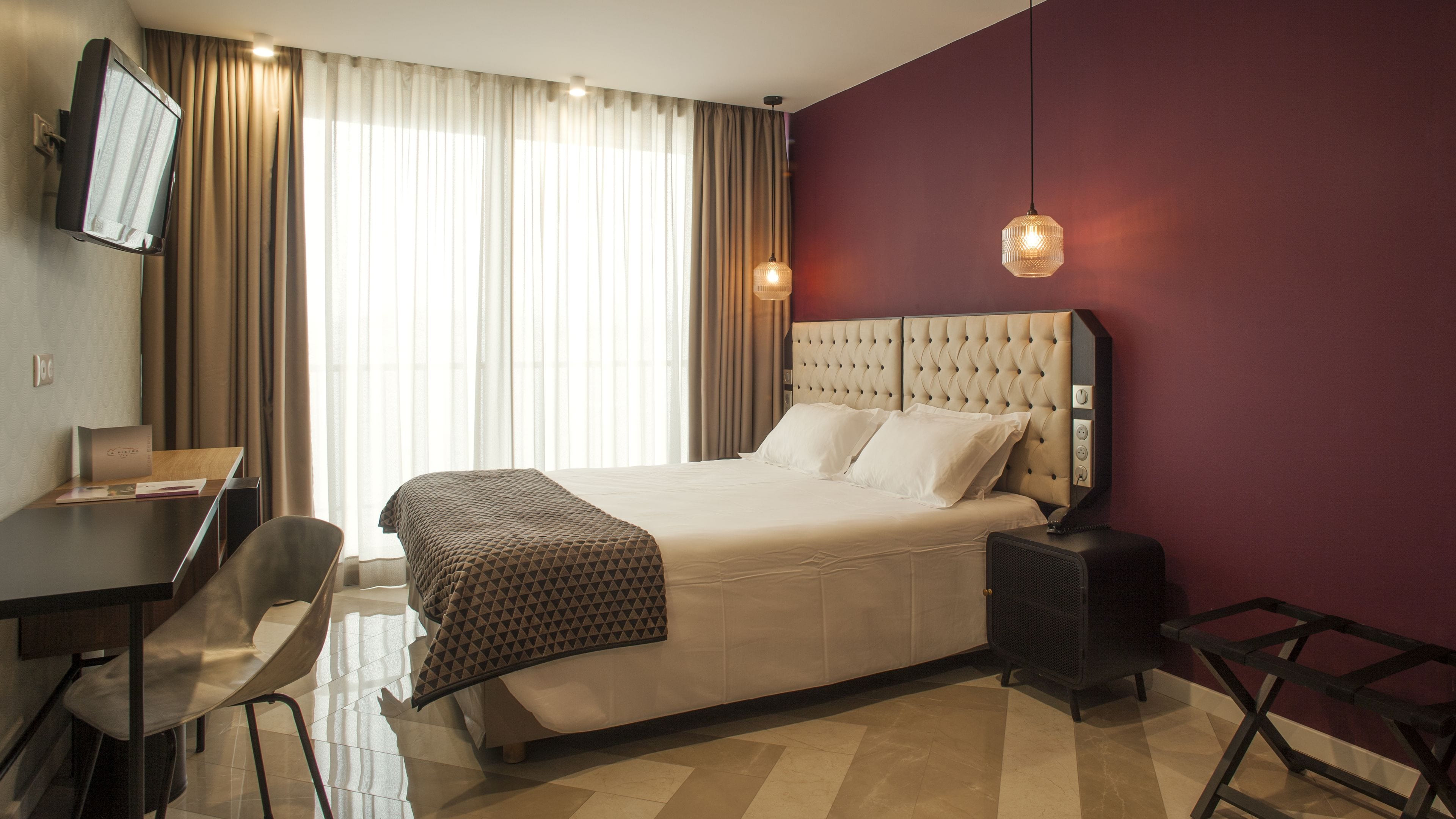 A hotel room with a bed, a desk, a chair, a TV, and a window with curtains.
