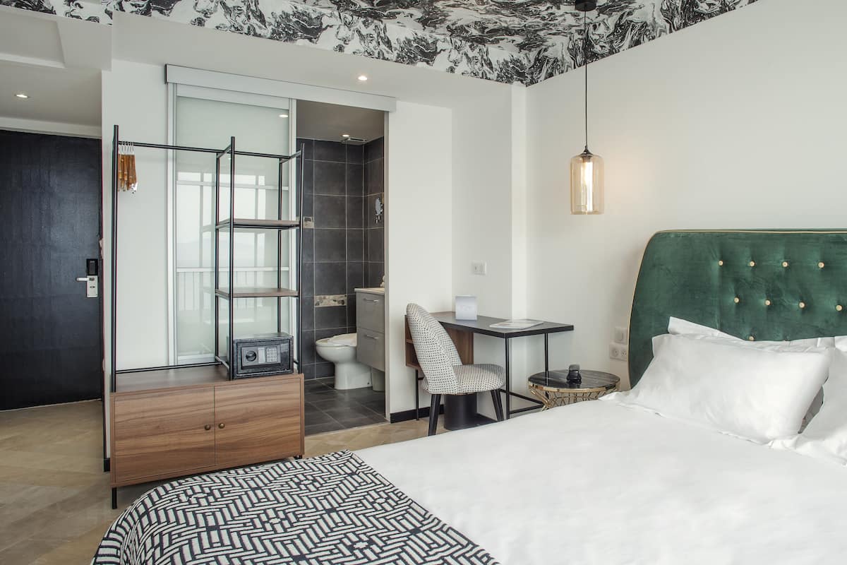 Une chambre d’hôtel moderne avec un grand lit, un bureau et une chaise. On peut apercevoir la salle de bain à travers une porte ouverte.