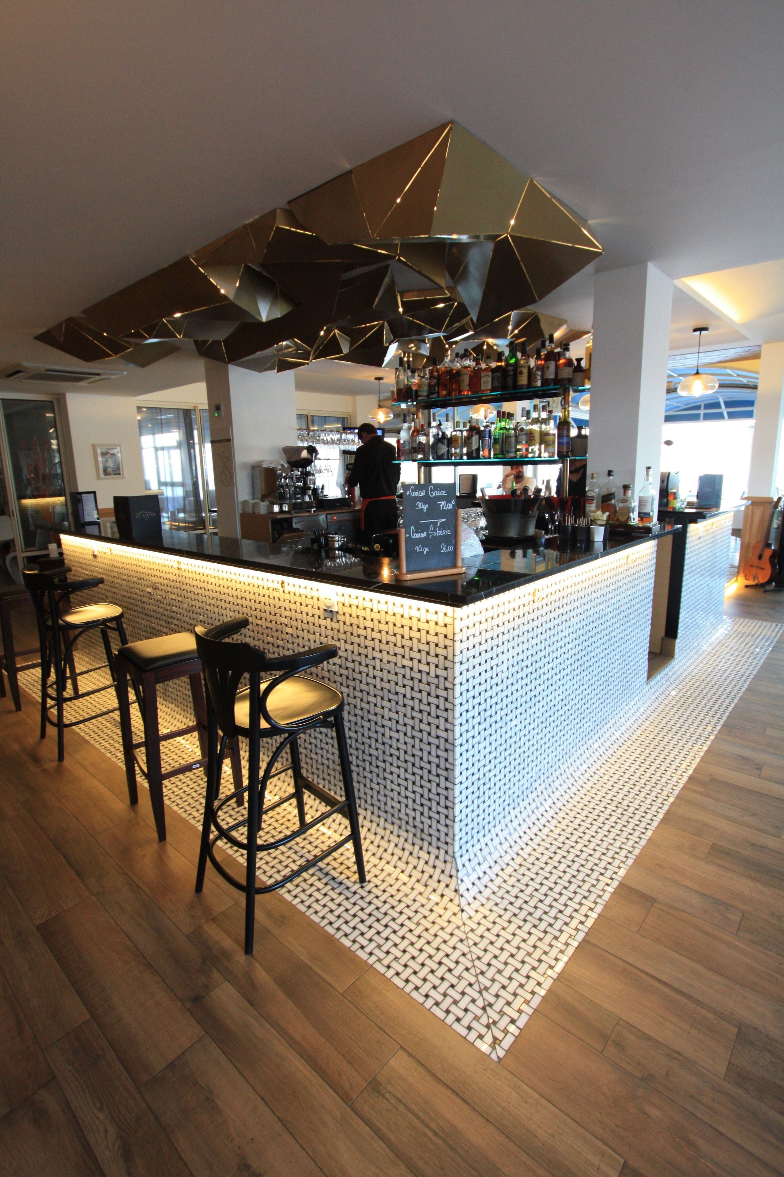 A modern bar area with a geometric ceiling design, a bar counter, and multiple stools.