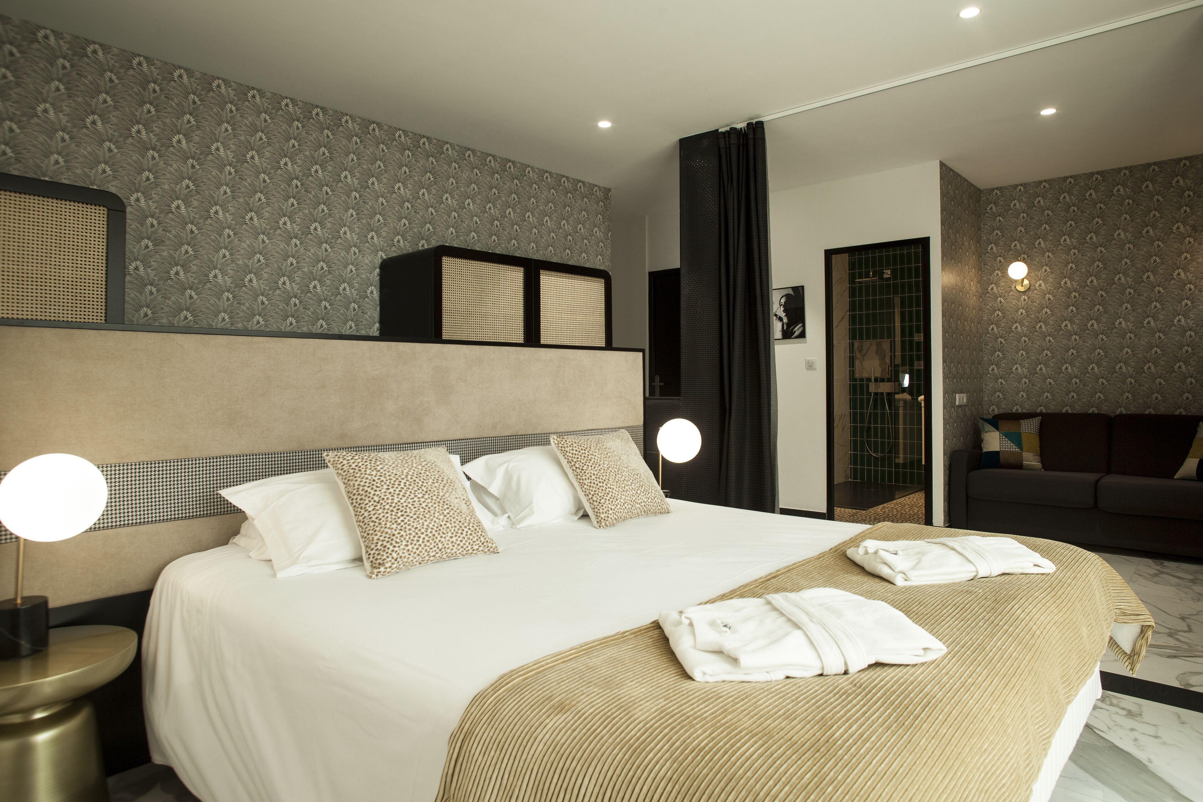 A modern hotel room with a large bed, two bedside lamps, a sofa, and a bathroom visible through an open door.