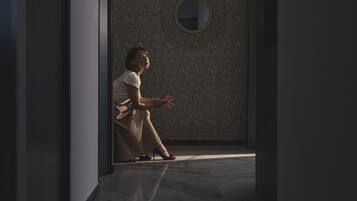 A person sitting by a window with a patterned wall and a round mirror.