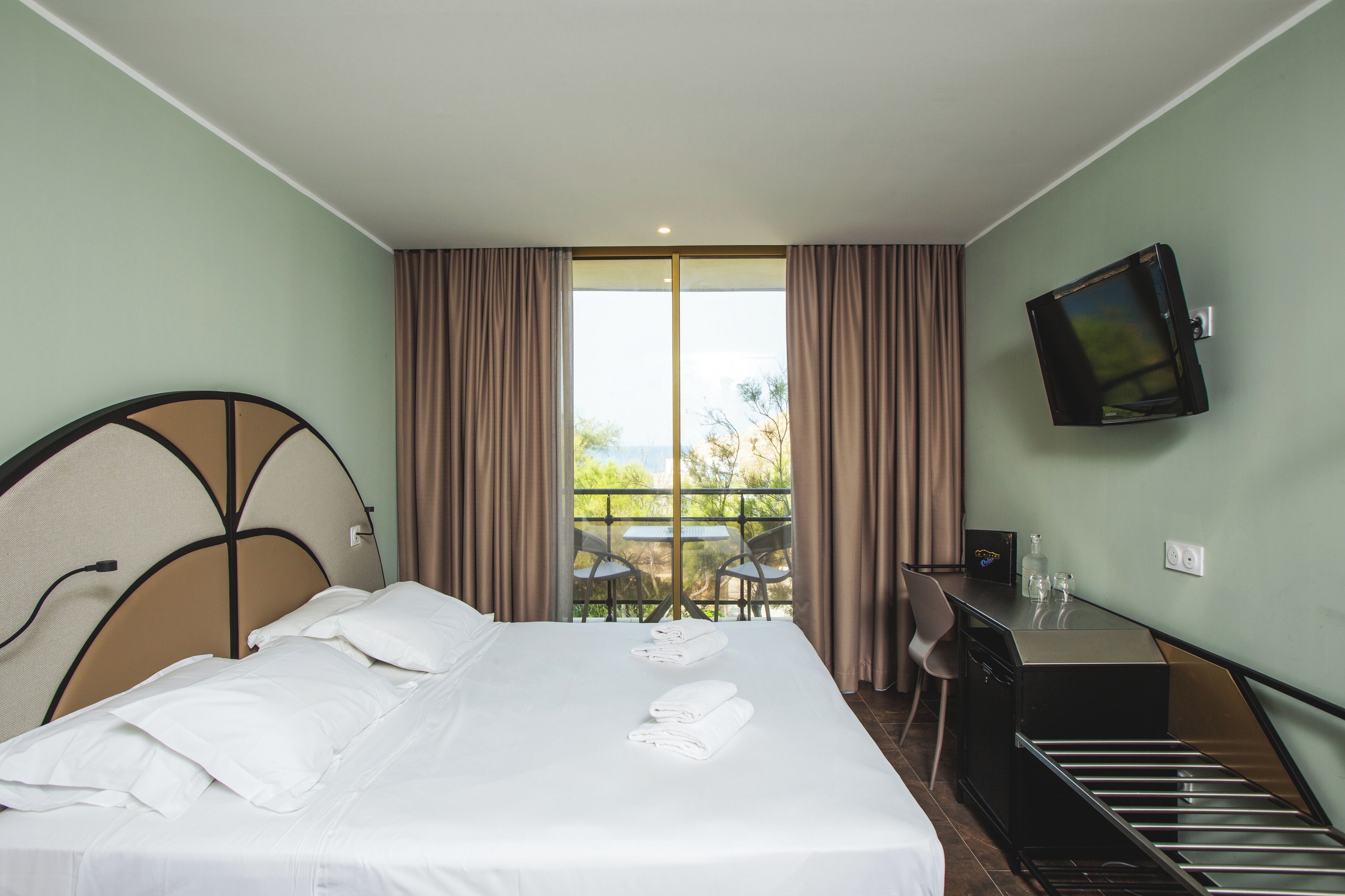 A hotel room with a bed, a desk, a TV, and a balcony with chairs.