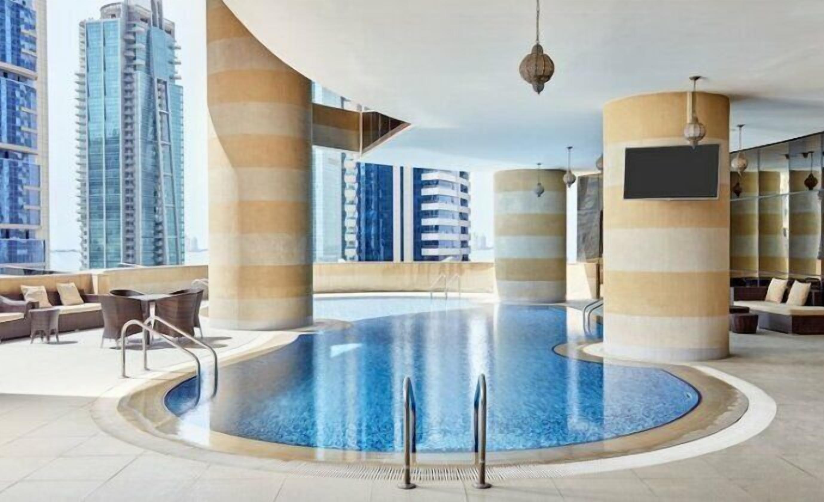Une piscine intérieure offrant une vue sur de hauts immeubles, entourée d’espaces de détente et d’une grande colonne.