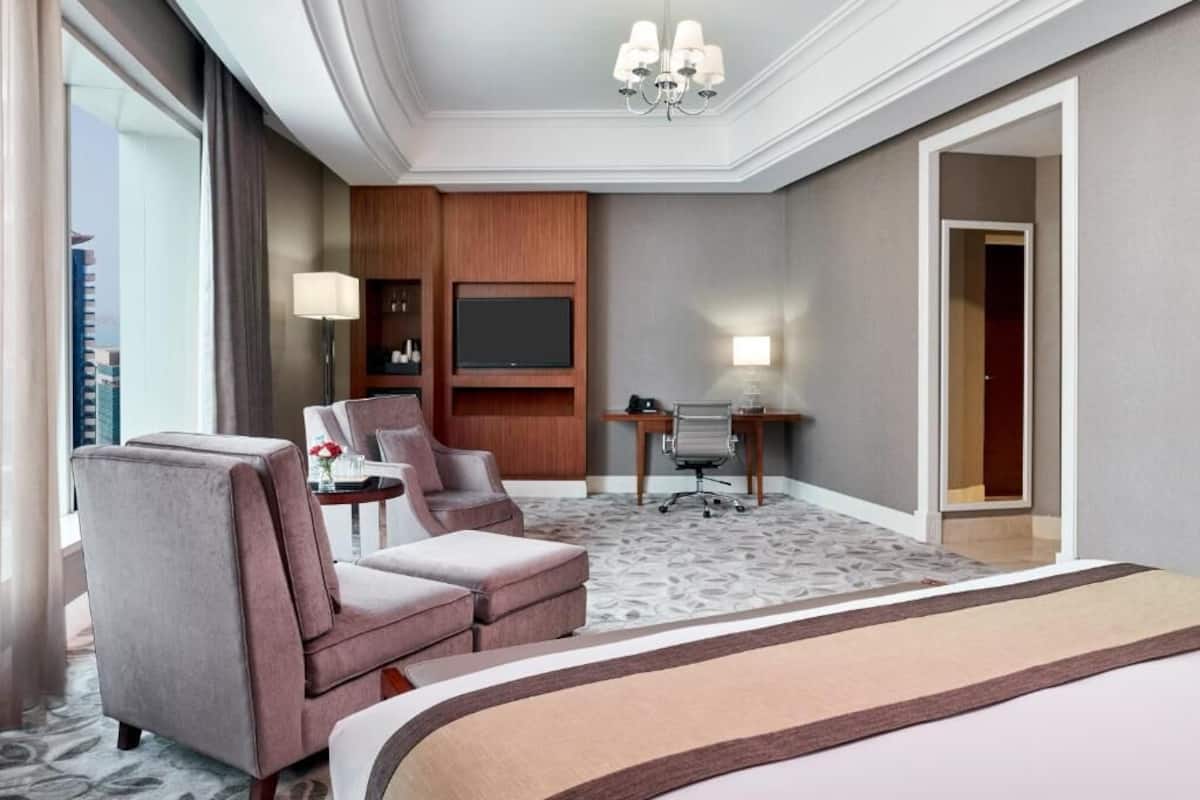Une chambre d’hôtel avec un grand lit, deux fauteuils, un bureau avec un ordinateur, un téléviseur à écran plat et une grande fenêtre.