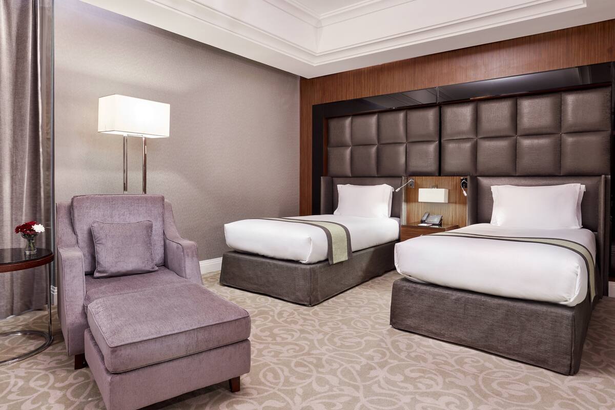 Une chambre d’hôtel avec deux lits, une chaise longue, une table de chevet avec une lampe et une moquette à motifs.