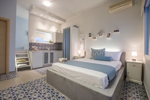 A compact bedroom with a bed, bedside tables, a small desk, and a kitchenette. - Aphrodite Studios (Astypalaia)