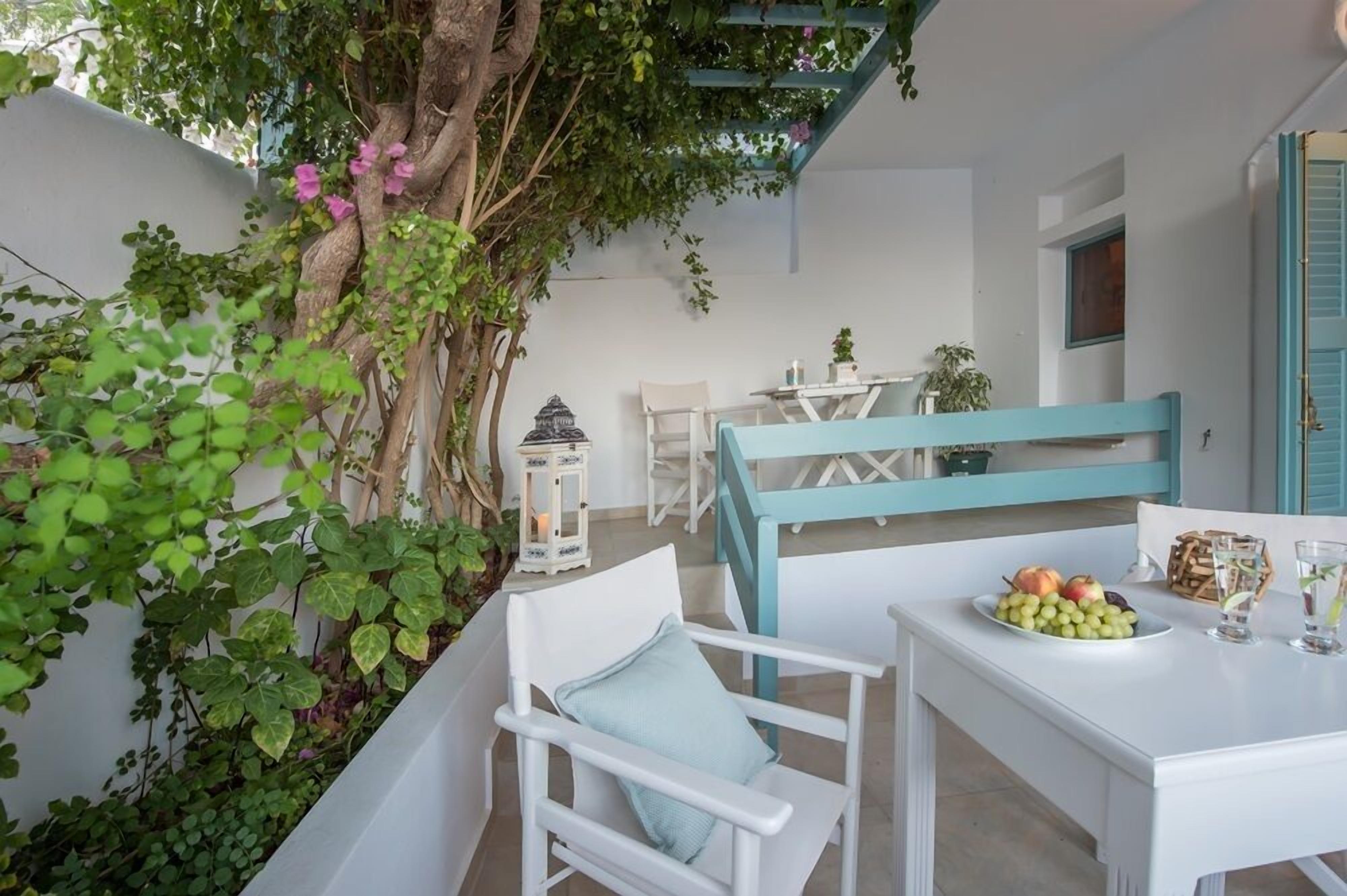 Un espacio al aire libre con mobiliario blanco, una mesa y sillas, rodeado de follaje y un árbol.