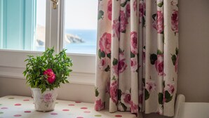 A window with a view of the sea, a potted plant with pink flowers, and curtains with a floral pattern. - Aphrodite Studios (Astypalaia)