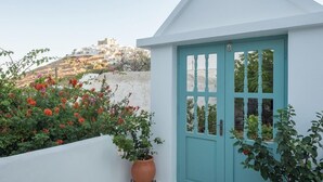 A white building with a turquoise door, a potted plant, and a view of a hillside with buildings. - Aphrodite Studios (Astypalaia)