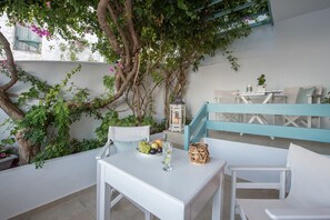 A white outdoor seating area with a table set for two, surrounded by greenery and a tree. - Aphrodite Studios (Astypalaia)