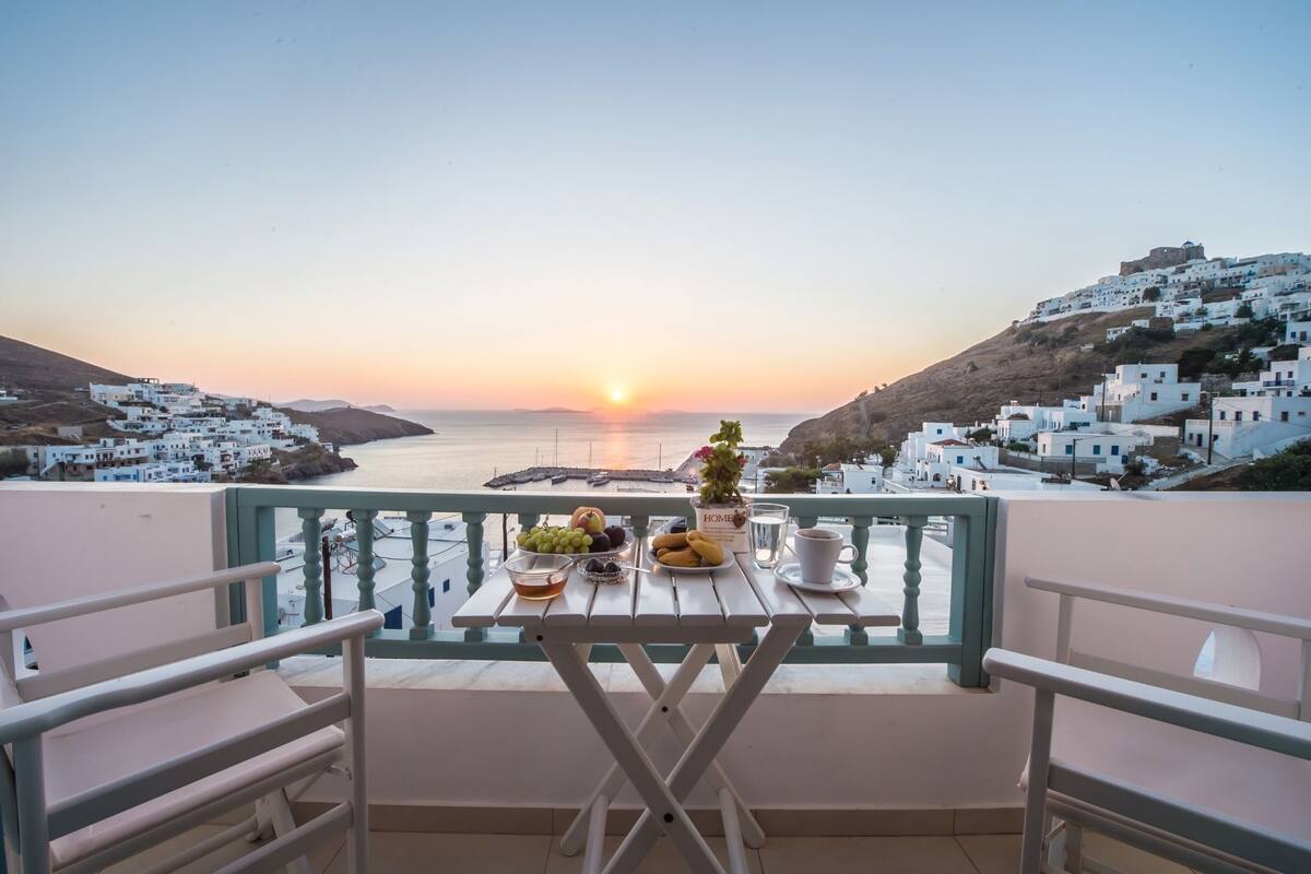 A balcony with a table set for breakfast overlooking a coastal village at sunset.