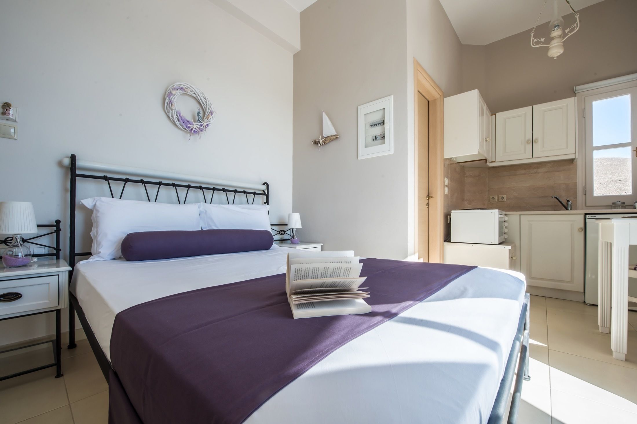A neatly made bed with a purple blanket and a stack of books, a bedside table with a lamp, a small decorative wreath on the wall, and a view of a kitchen area with white cabinets and a window.