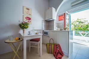 A compact kitchen with white cabinets, a microwave, and a small dining table with a red chair. A colorful bag is placed on the floor next to the table. - Aphrodite Studios (Astypalaia)