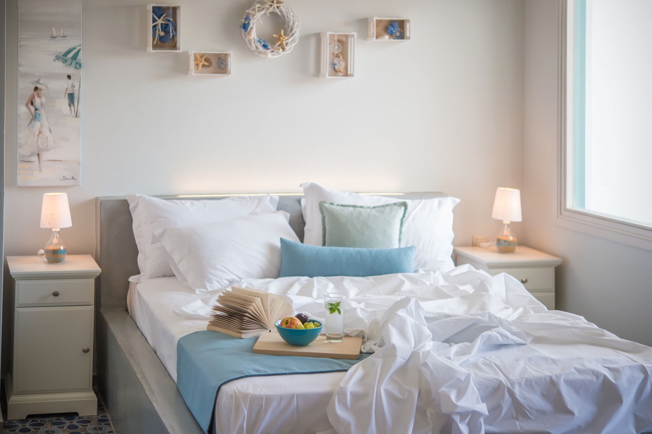 A neatly made bed with white and blue bedding, a bowl of fruit, and a glass of water on a bedside table.