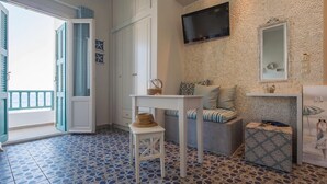 A room with a patterned floor, a sofa with cushions, a small table with a basket, a television mounted on a textured wall, and a mirror above a cabinet. - Aphrodite Studios (Astypalaia)