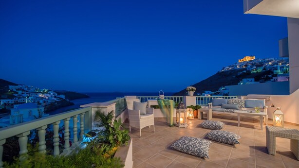 A terrace with outdoor seating, a view of the sea, and a hill with buildings. - Aphrodite Studios (Astypalaia)