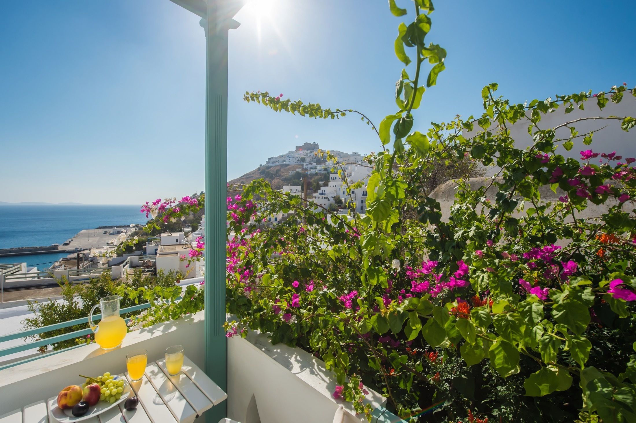A balcony with a table set for a meal, overlooking a coastal town and the sea.