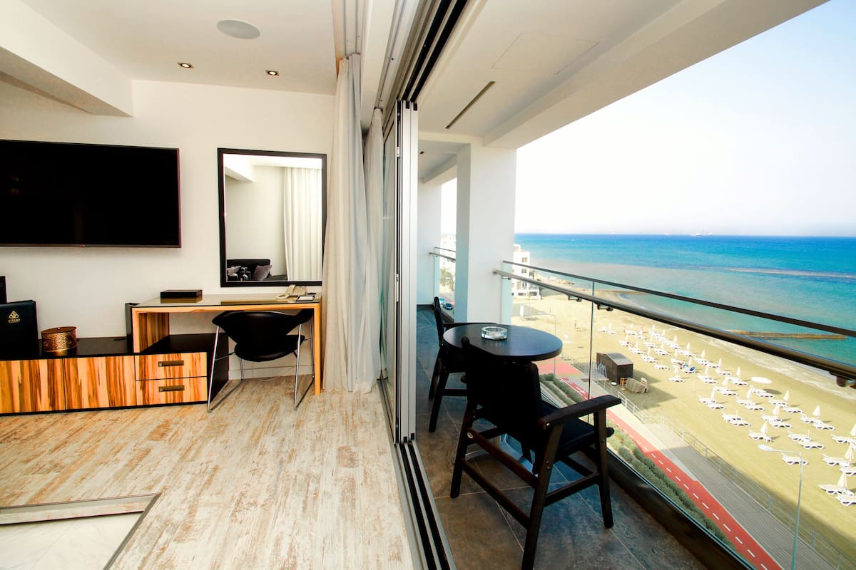 Une chambre d’hôtel moderne dotée d’une grande fenêtre donnant sur une plage. La chambre comprend un bureau, une chaise et un téléviseur à écran plat.
