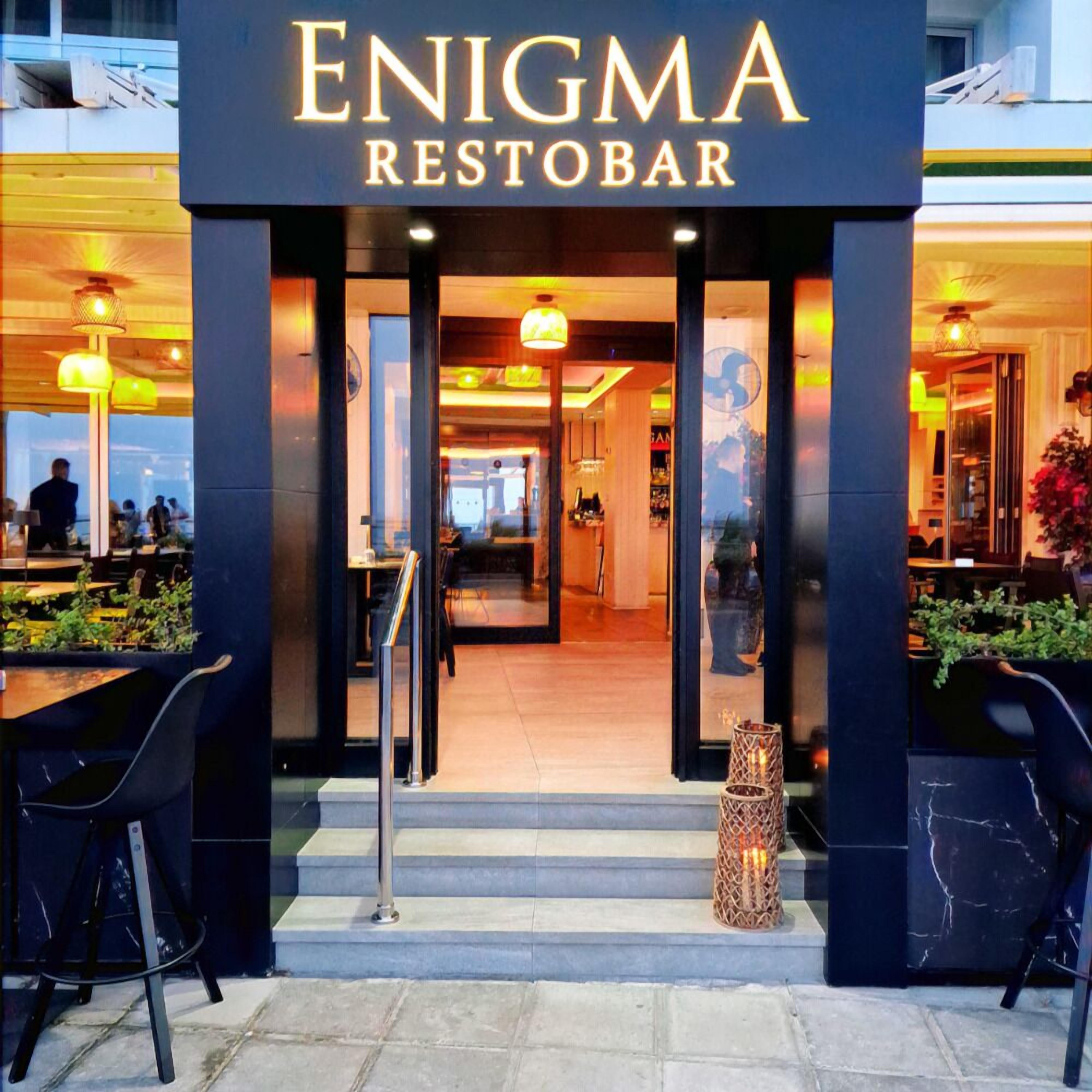 The entrance to Enigma Restobar with a modern design, featuring a glass door, outdoor seating area, and a visible interior with tables and chairs.