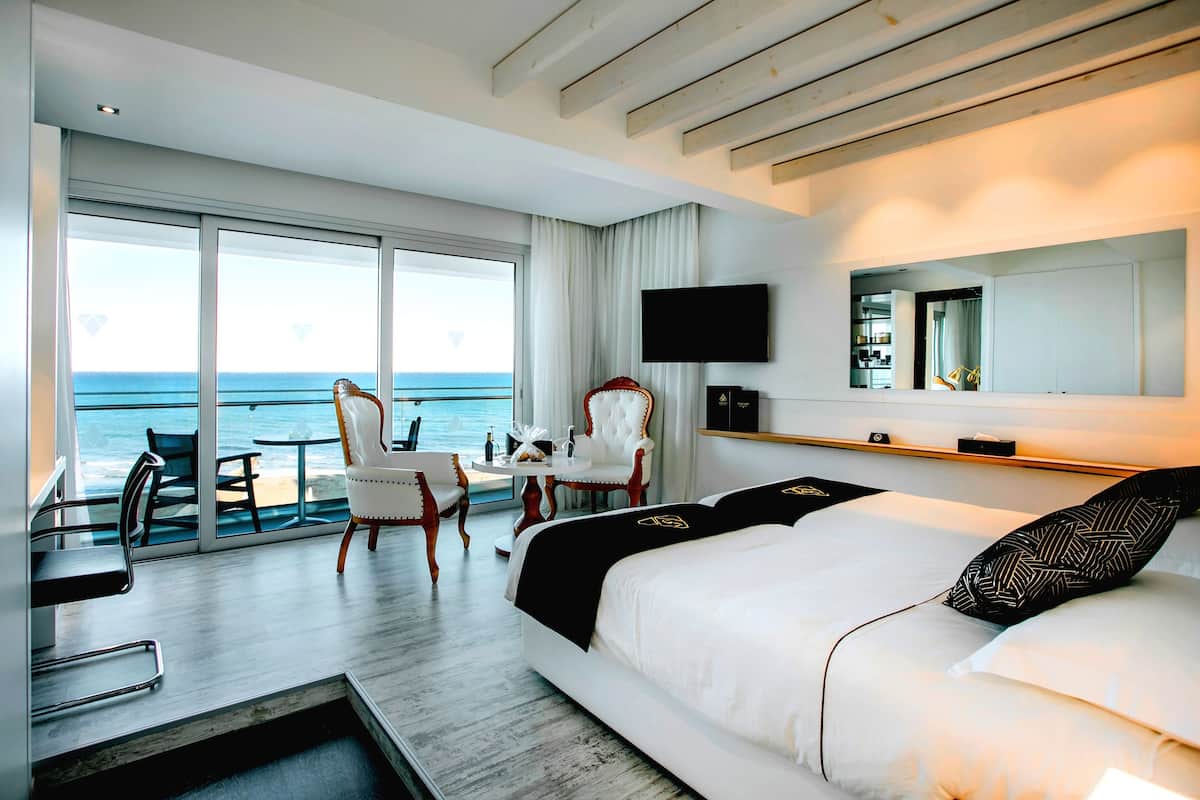 Une chambre d’hôtel moderne avec un grand lit, un bureau et une chaise. Un téléviseur est fixé au mur et de grandes fenêtres offrent une vue sur l’océan.