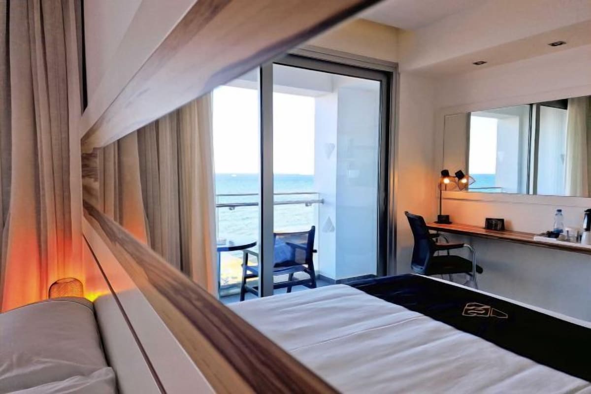 A hotel room with a bed, a desk, a chair, a large window, and a view of the sea.