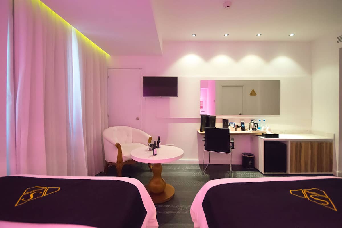 Une chambre d’hôtel avec deux lits, une petite table ronde, une chaise blanche, un bureau avec un ordinateur, un miroir et un téléviseur fixé au mur.